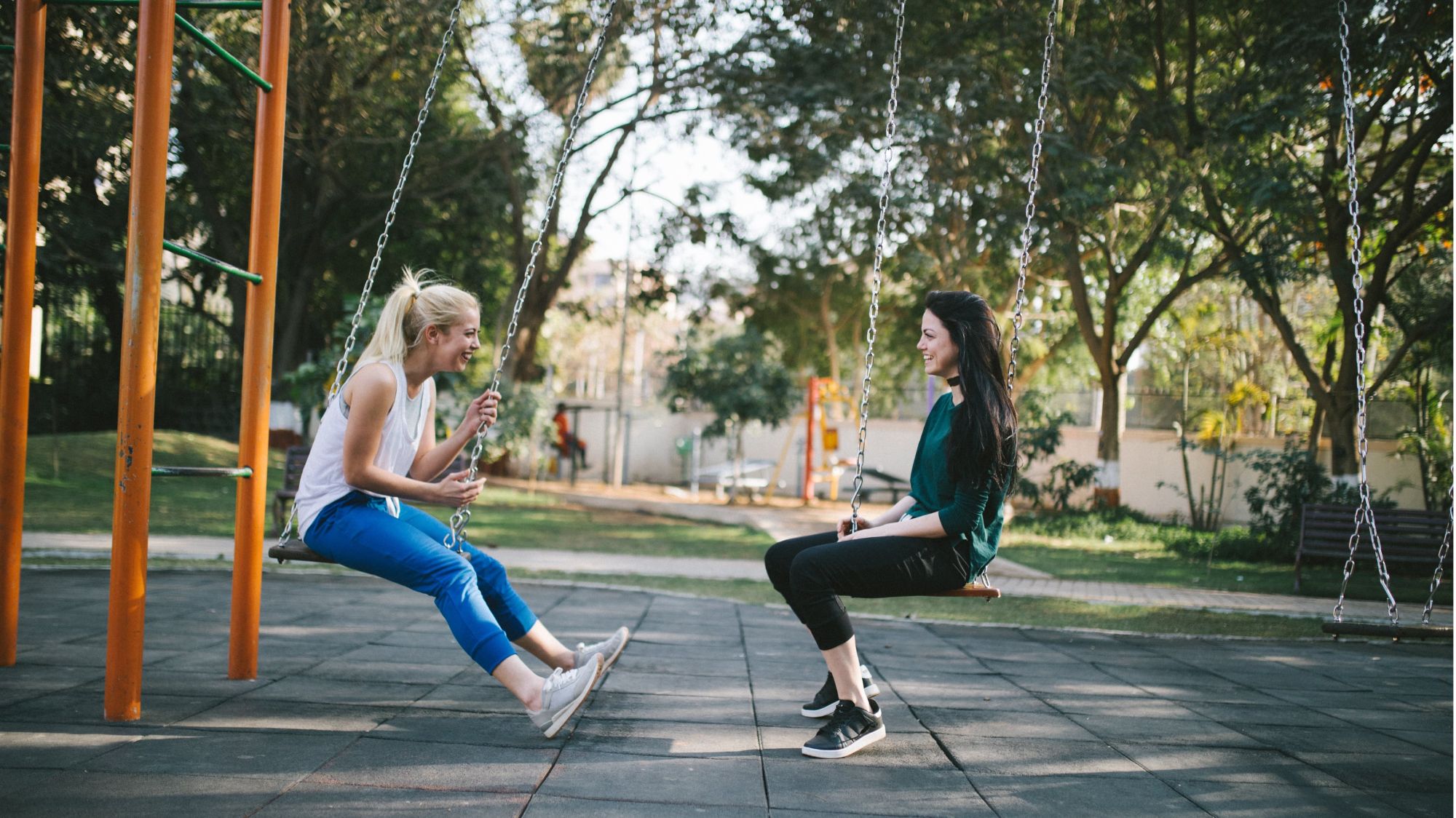 two teenage girls, sitting on swings in park, facing each other, smiling and conversing with one another with large trees, bench, and younger kid park in background.