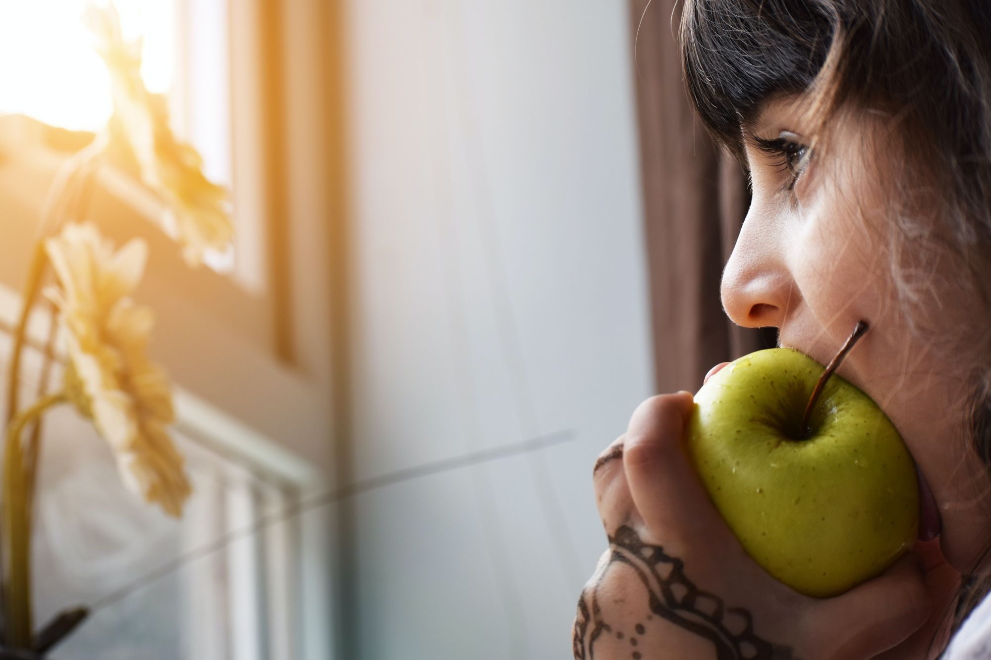 A side profile picture of a young girl, about 8 or 9 years old, biting into a green apple with mehndi on the hand grasping the apple. She is staring out of a window, with two or three sunflowers on the window sill in front of her with sunshine peering through the bottom corner of the top panel of the horizontally divided window.