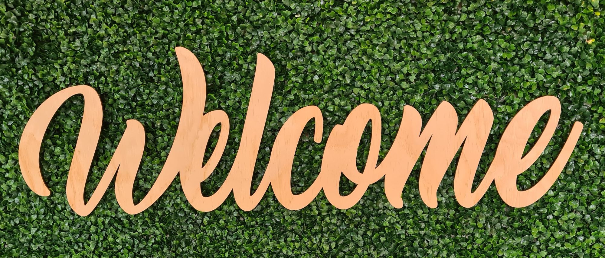 A cursive "Welcome" sign made of wood, sitting on top of green plants, providing a contrast between the maple-looking wood and green manicured plants.
