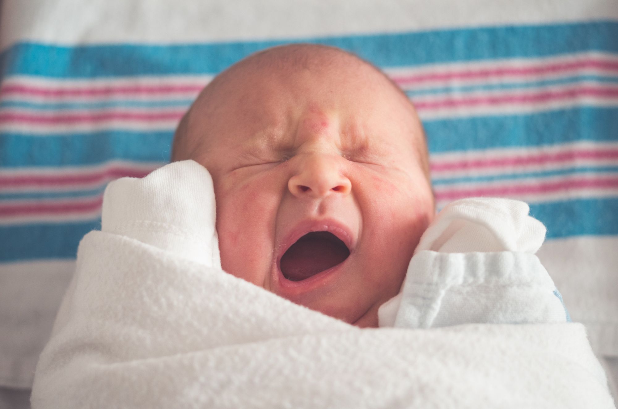 A newborn baby yawning while swaddled in a blanket. Contrasted with a teal and pink striped newborn blanket in background.